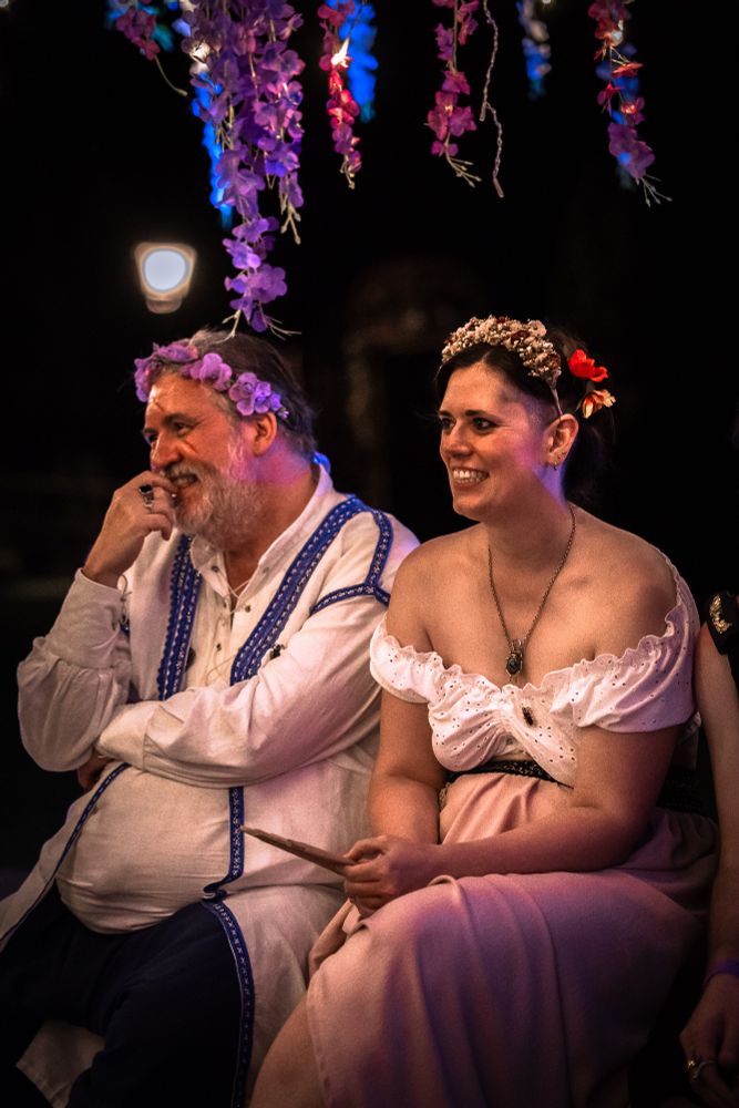 Su Seguro Servidor con traje de inspiración medieval y corona de flores, sentado junto a una mujer con estilo similar, bajo unas guirnaldas de flores. Ambos muy sonrientes. Es de noche.