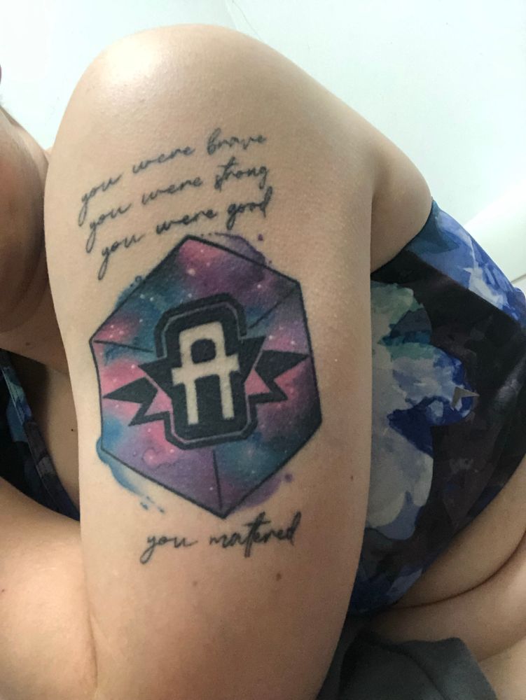 The letter A over a galaxy filled morphing cube, with the words “You were strong, you were brave, you were good. You mattered.” on a arm tattoo
