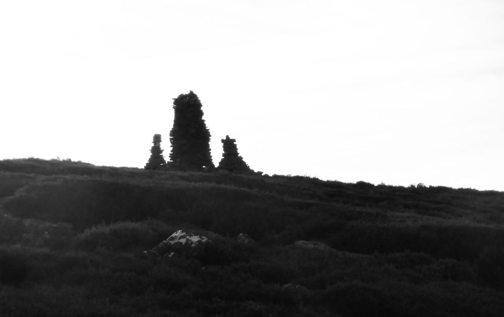 Black and white silhouette of a moorland skyline with  thee rough stone cairns, the central one higher than the other two.