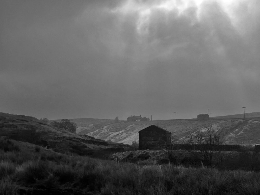 Black and white image. The gable of an isolated fell barn stands in rolling hills of heather and sedge with the odd clump of bare trees. A deep valley cuts the landscape, on the other side of the valley is a ridge with a farmhouse and a barn connected by a series of telegraph poles. The sky above is heavy black cloud but there’s a momentary break to allow silver rays to show.