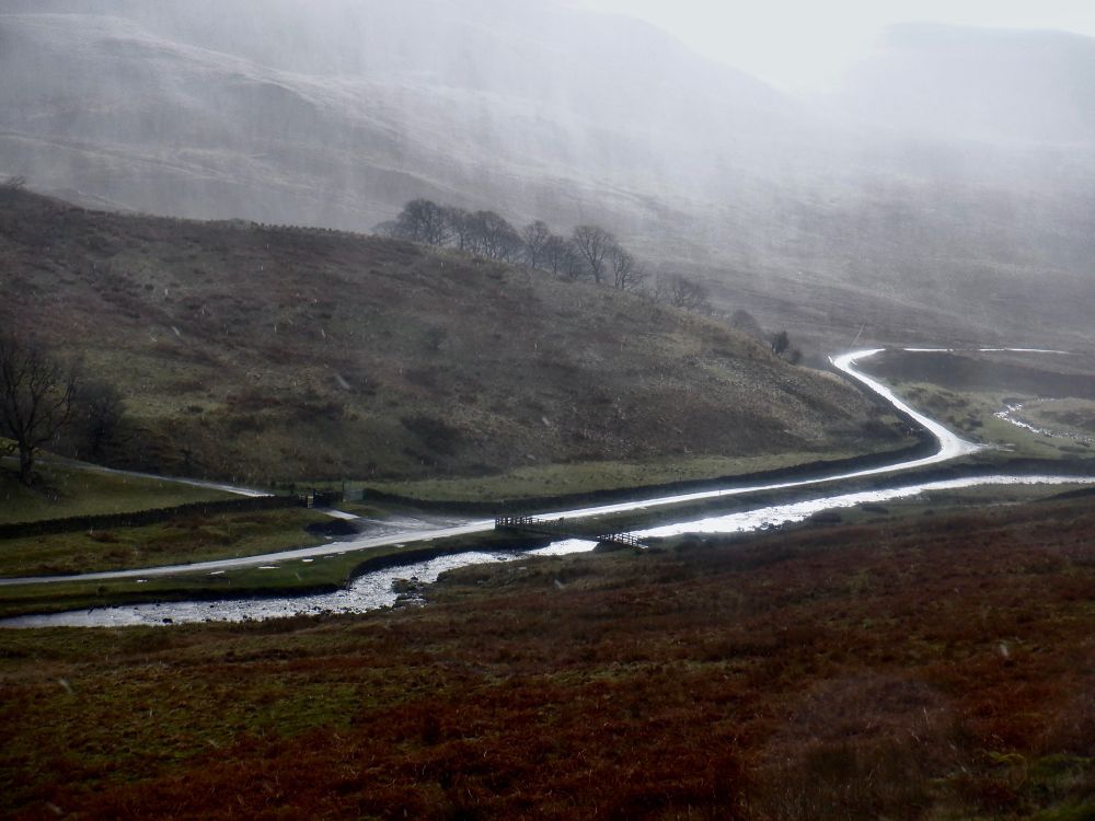 A road and a river, silver lit by a fleeting sun, pass through a valley in the hills. A few trees stand on a ridge running down into the valley, partly obscured by pulses of grey rain.