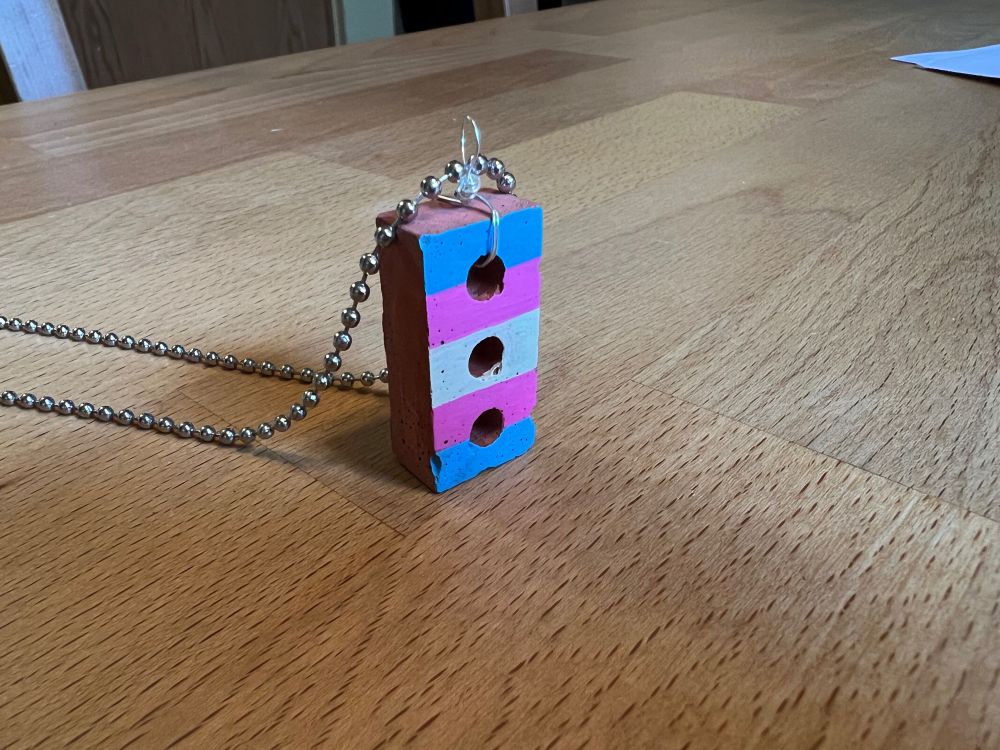 Photo of a small trans pride charm, a brick painted with blue, pink, and white stripes, on a thin chain