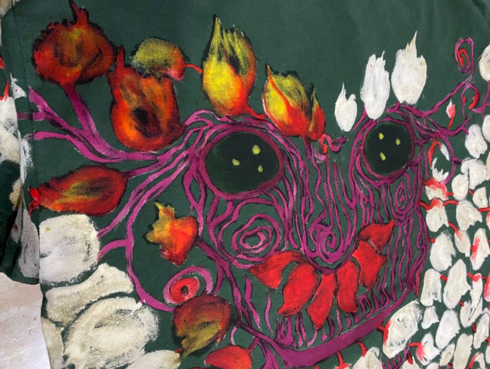 Close-up view of the face of a blackberry spirit in the process of being painted, its wooden mask rendered with deep maroon lines and its leaves golden and red.