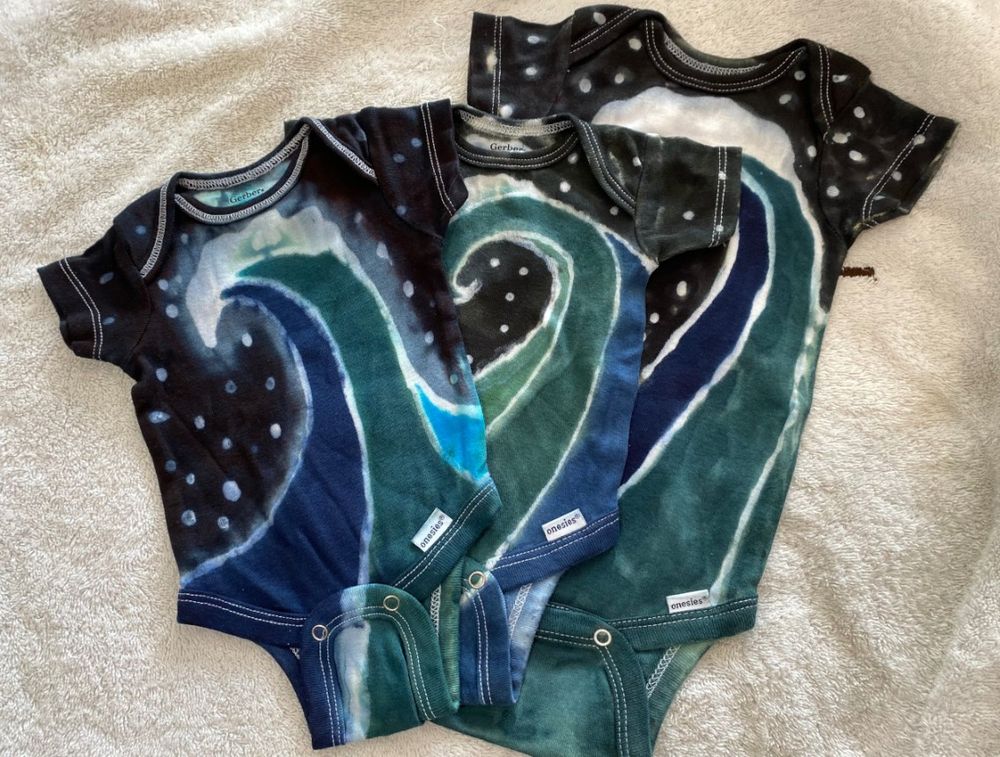 A pair of hand-painted and hand-dyed baby onesies featuring a Night Ocean pattern of a cresting wave against the night sky.
