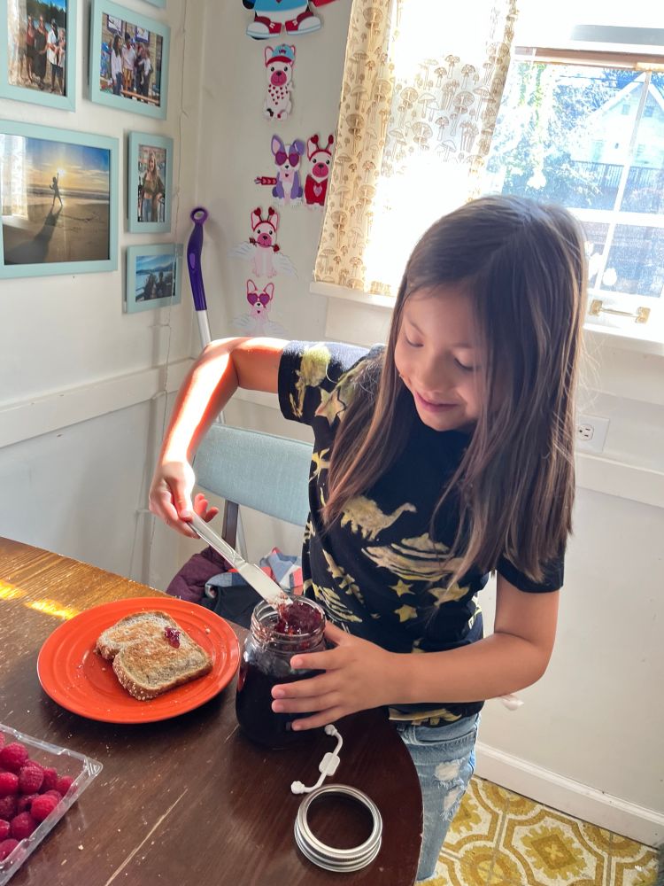 An adorable child using a butter knife to get grape jelly out of a giant jar to put onto a slice of bread with peanut butter.