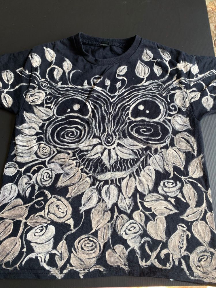 The front of a hand-painted T-shirt, size M, featuring a cheerful Green Man face surrounded by leaves, thorny vines and roses.