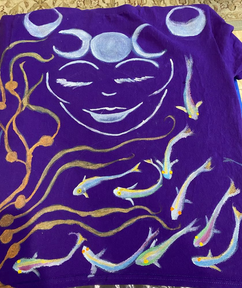The front of a hand-painted T-shirt, featuring the serene face of a Moon Goddess with waving undersea kelp and prismatic silvery Moon Fish.