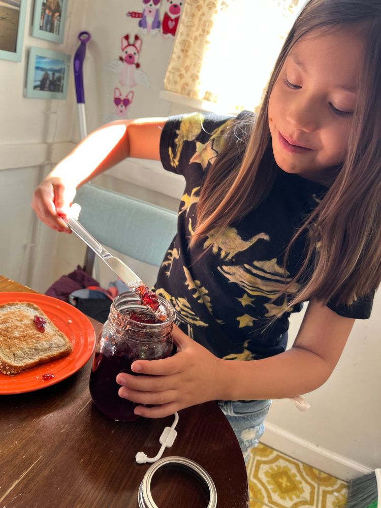 An adorable child using a butter knife to get grape jelly out of a giant jar to put onto a slice of bread with peanut butter.