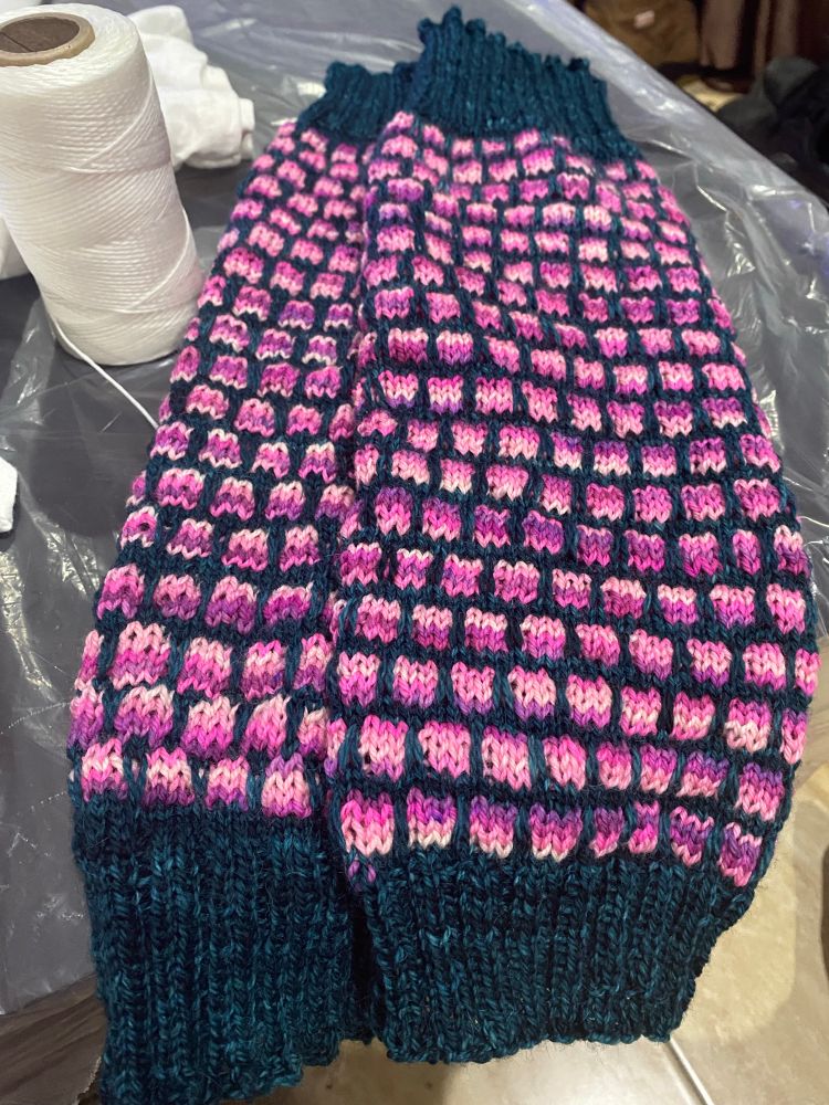 A new pair of hand-knitted leg warmers made from two hand-dyed yarns: a deep green-teal as the background, and a pattern of squares made from a yarn that shifts through a range of bright, cheerful pink and lavender tones.