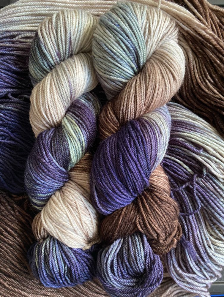 A photo of Slan Baby’s “Elisabeth’s Grey” colorway, four skeins of hand-dyed yarn that incorporate dark and golden brown with silver-blue-grey. The base in this photo is Simple DK, a superwash yarn made from 75% merino wool and 25% nylon.