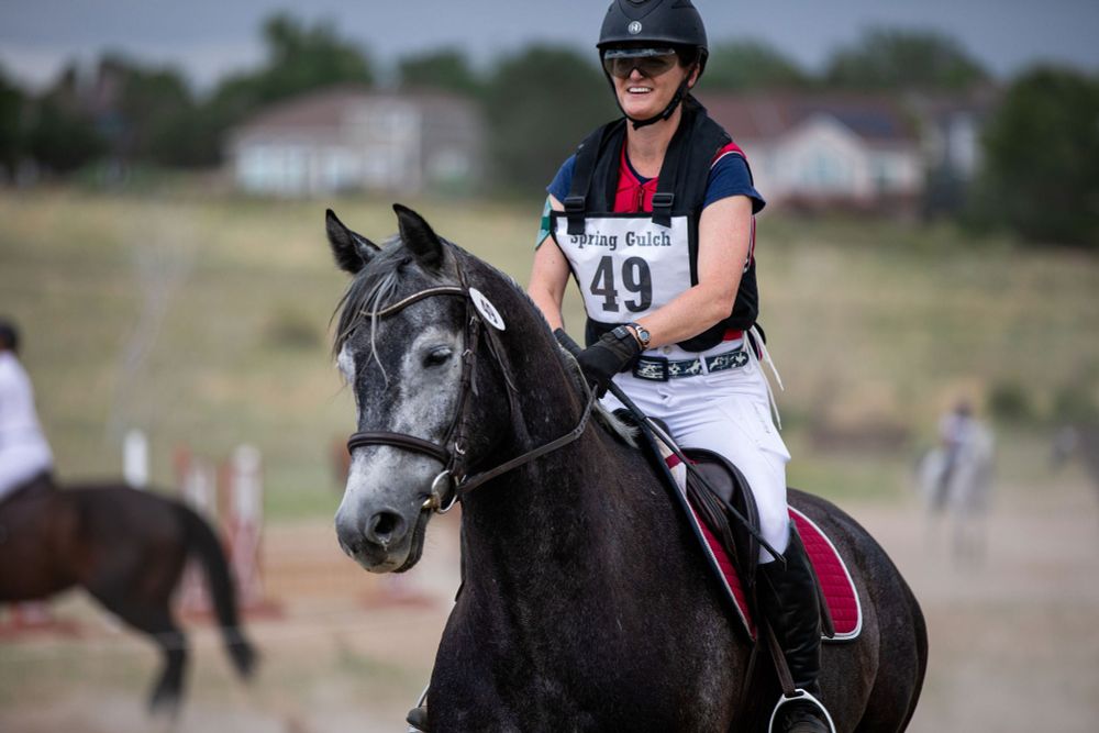 Picture of me riding my young horse, Phantom, at an eventing competition. Phantom is a dark grey Oldenburg horse with circles of lighter hair around his eyes. I am a middle aged woman sitting on his back, wearing a short sleeve navy shirt and red protective vest with my competition number pinny over the top. The number is 49. I'm wearing white riding breeches and black tall boots, sitting in an english saddle with a bright maroon saddle pad that matches my protective vest. In my left hand, I'm holding a riding crop. I am smiling and have a helmet and sunglasses on my head. 