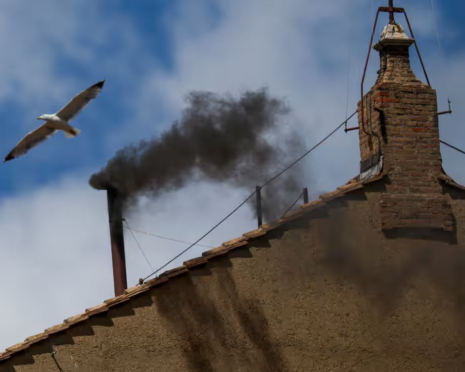 This image shows the roof of a rustic building with a tall chimney made of old bricks. From the chimney, thick black smoke is billowing out into the sky, forming a heavy, dark cloud. The smoke contrasts sharply with the bright blue sky and scattered white clouds. A seagull is captured mid-flight in the upper left portion of the image, wings spread wide as it glides near the rising smoke. The structure has a weathered, textured appearance, and several wires are anchored to the chimney, possibly for stability. The overall atmosphere suggests an old, possibly religious or historical building—this scene is notably reminiscent of the Vatican’s Sistine Chapel chimney during a papal conclave, where black smoke traditionally signifies no decision has been made in the selection of a new pope.
This image likely depicts a scene from the Vatican during a papal conclave—the secretive and highly symbolic process by which a new pope is elected by the College of Cardinals.

The chimney in the photo is attached to the Sistine Chapel, the building where the cardinals meet to vote. The black smoke rising from the chimney is a significant signal: it means that a vote has been taken, but no candidate has yet received the required two-thirds majority to be elected pope. The smoke is created by burning the ballots along with chemical additives that ensure the color is clearly visible from outside.

This signaling process has been used for centuries. Black smoke (“fumata nera” in Italian) means no pope has been elected. When a pope is elected, the smoke that rises is white (“fumata bianca”), announcing joyful news to the crowd gathered in St. Peter’s Square and to the world.

The inclusion of a seagull in the image adds an accidental but often noticed detail—birds, especially seagulls, are sometimes seen perching on the chimney, and this has been playfully interpreted by the media and observers as symbolic or simply amusing.