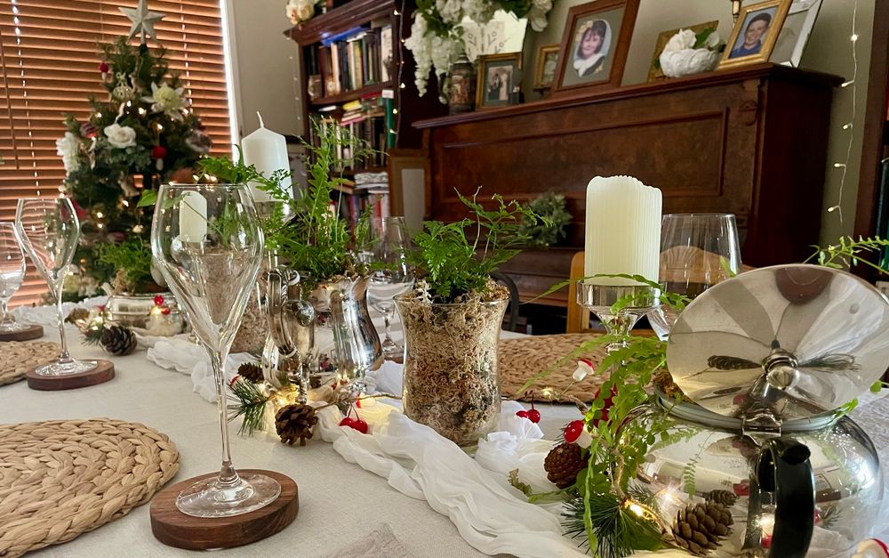 Table dressed with white muslin runner, candles, strings of fairy lights with sprigs of pine, berries & toadstools and various ferns in vintage silver teapots 