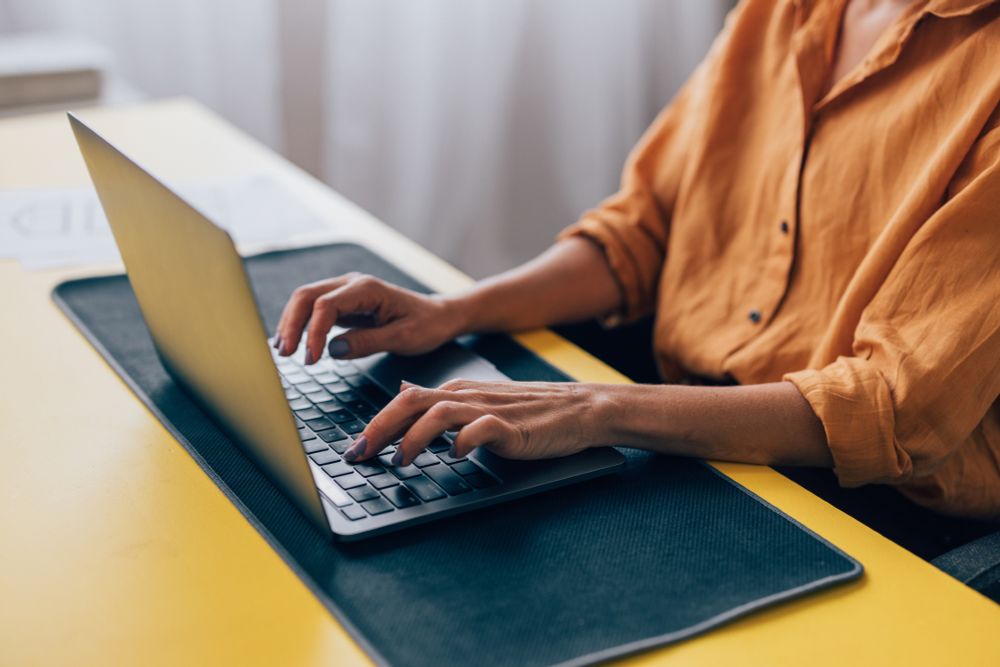 Close-up of a woman in a yellow shirt typing on a laptop, situated at a vibrant workspace with paperwork around.