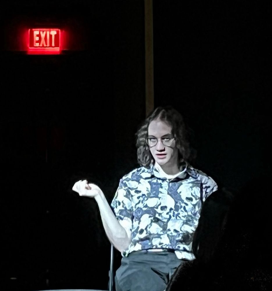Levi sits under a spotlight and gazes at the camera. A red exit sign is visible in the dark behind him. This image is from a staged performance.