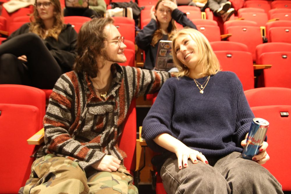 Levi is sitting in the audience of a theatre. He has his arm around his friend Anya as they smile at one another.