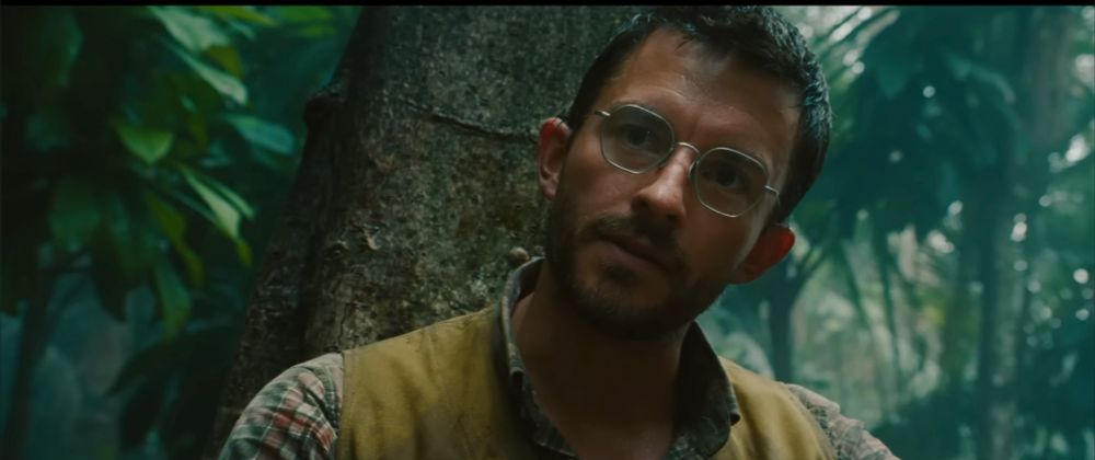 Jonathan Bailey looks just outside of frame. He wears glasses, and a plaid shirt with a yellow vest. Tropical greenery fills the space behind him. 