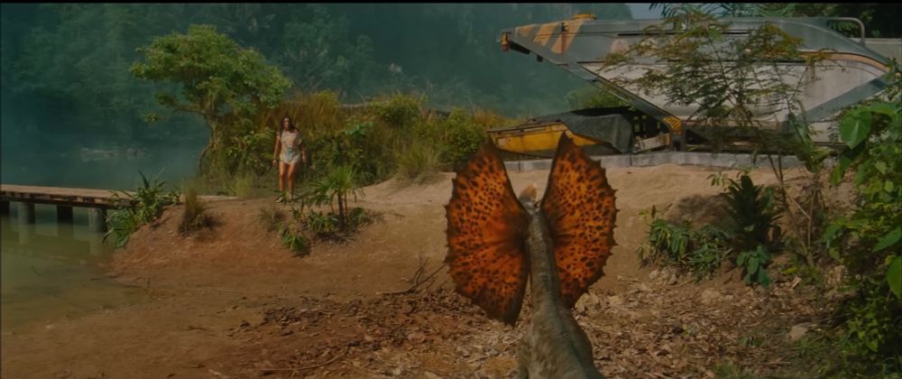 A small dinosaur with an extended, orange frill covered in black spots faces a woman in shorts and a tank top. A woman stands near a lake and a futuristic-looking aircraft, .The setting is a tropical jungle with a wooden dock leading into the water.