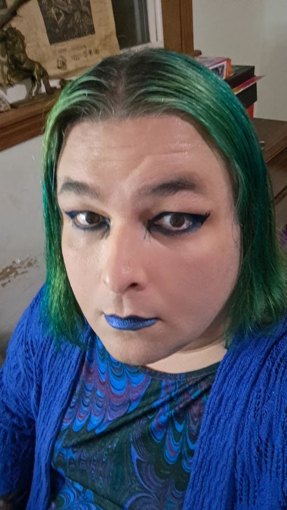 A forty-two year old woman with green hair, blue eyeshadow, black eyeliner, blue lipstick, a blue dress, and a blue cardigan. She is staring directly into the camera.
