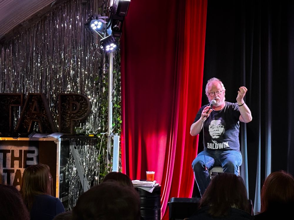 Robin Ince - a middle aged man with grey hair & a beard - sitting on a stage in front of red curtains. 