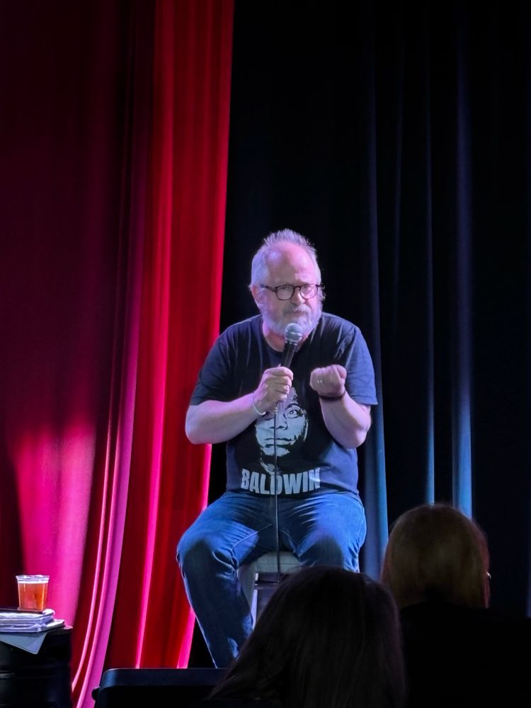 Robin Ince - a middle aged man with grey hair & a beard - sitting on a stage in front of red curtains 