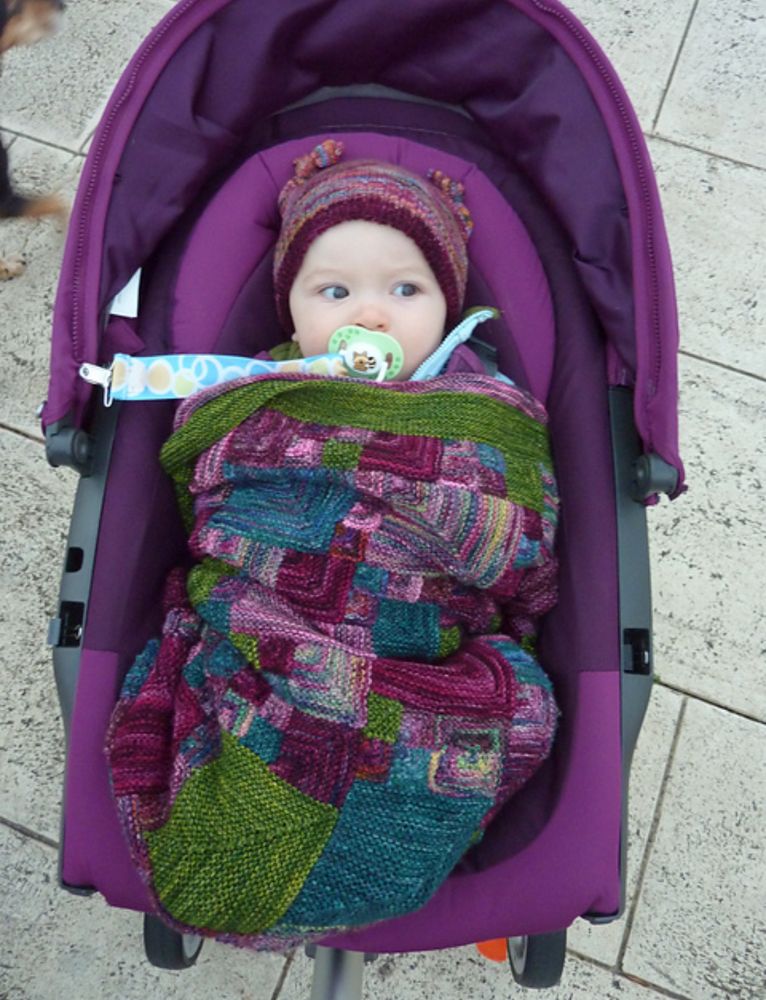 A baby giving side-eye in a purple pram, covered up with a multicolored hand knit blanket.