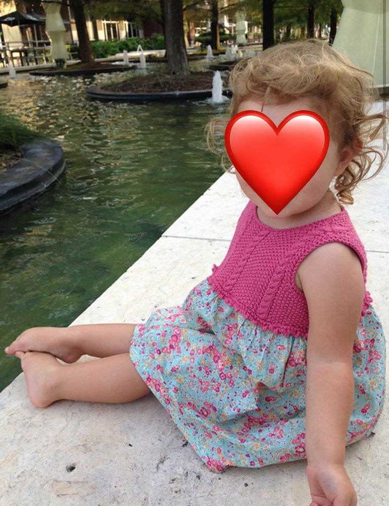 Little girl in a pink and blue hand knit and sewn dress, sitting on the ground beside a water feature. A red heart graphic over her face.