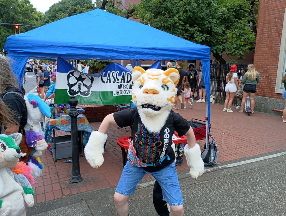 A photo of Kintsugi in partial standing in front of the Cascadia Furs tent at Portland Pride