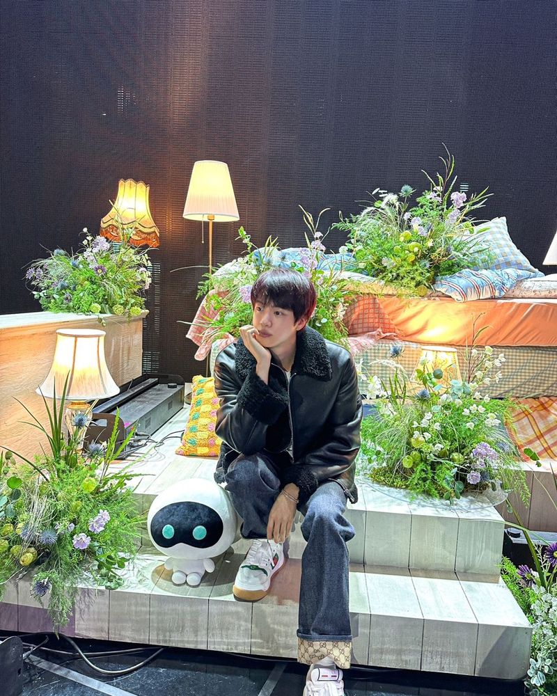 Jin of BTS is sitting on the steps of a small platform piled with mattresses, floral arrangements and a trio of lamps, with a stuffie of his alien astronaut character Wootteo by his side. Wootteo is white and has a small body with a large round head, a black mask and two round blue eyes.