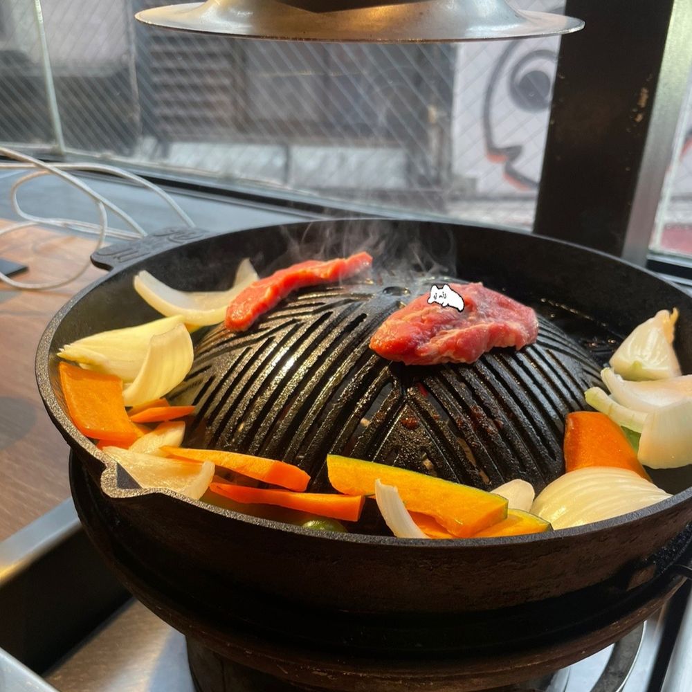 飲食店のジンギスカン用鍋の上で、羊肉、野菜(玉ねぎ、かぼちゃ、にんじんなど)が焼かれている写真 肉の上に泣きながら這いつくばるこいぬちゃんのスタンプが添えられている