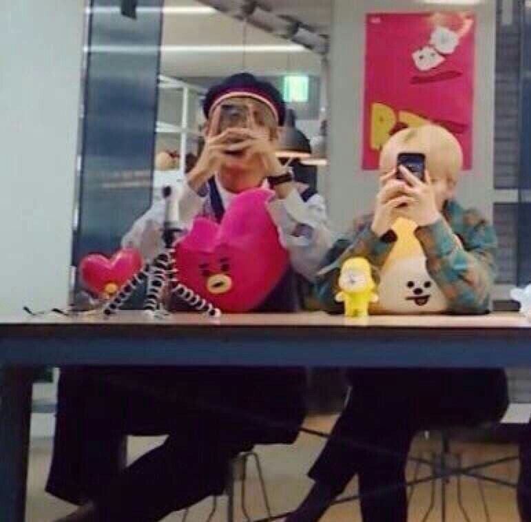 Taehyung and Jimin sitting at a table next to each other, looking at their phones. Jimin looks exceptionally tiny.