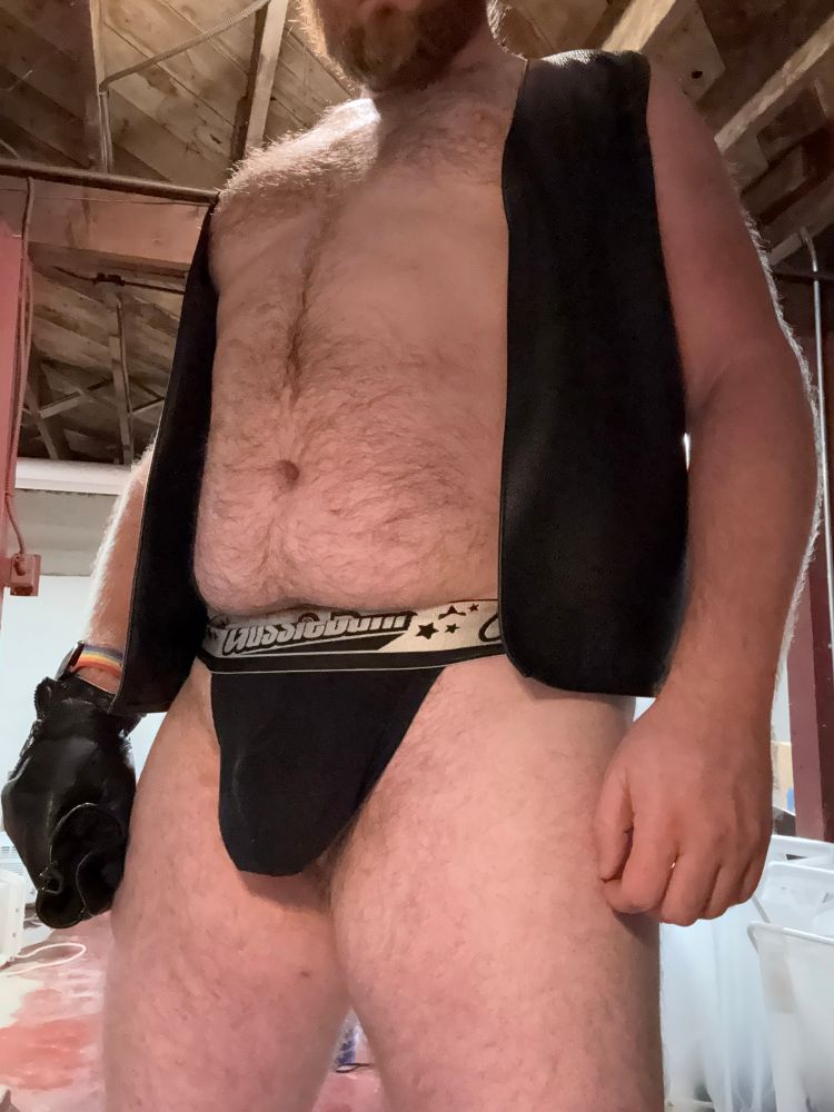 Man in a leather vest and a black jock, holding leather gloves. 