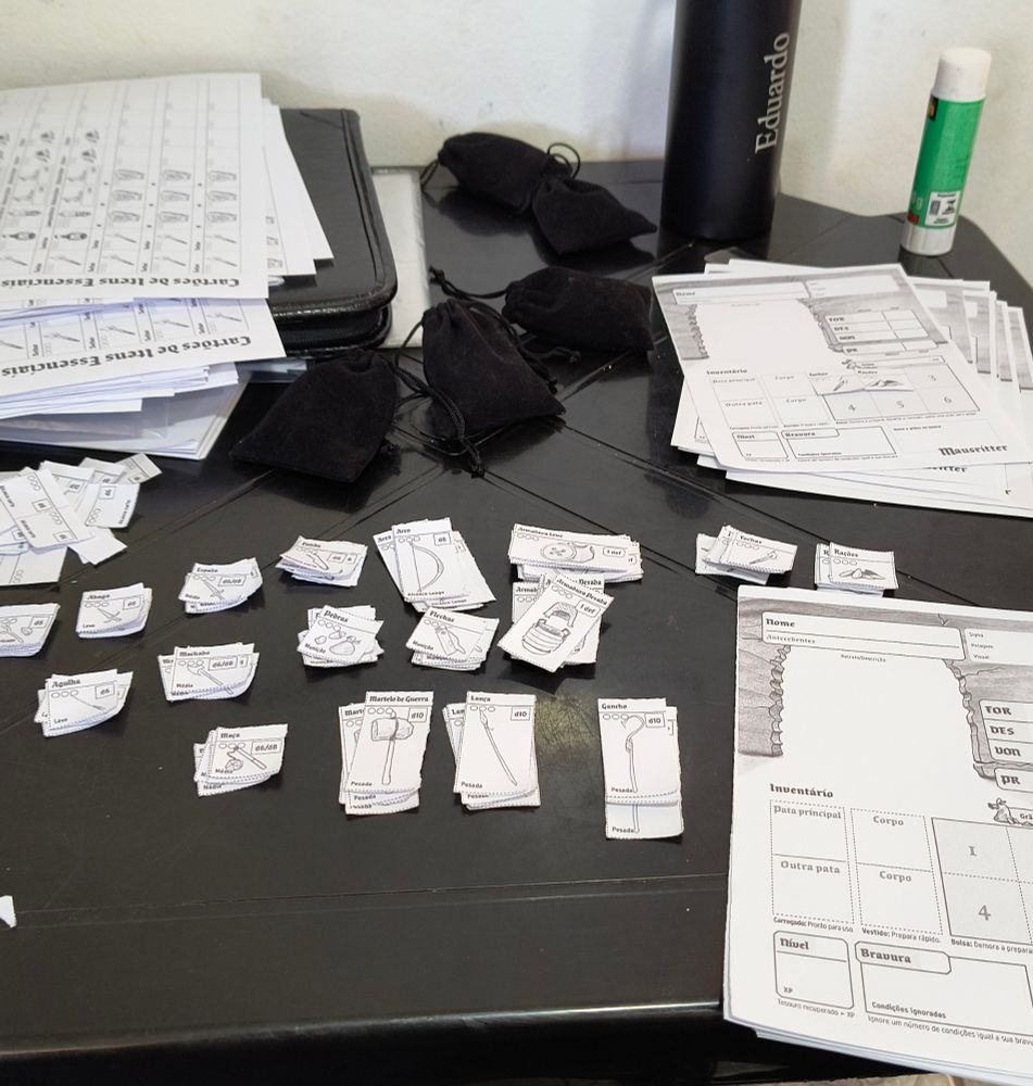 Pilhas de fichas de personagem, cartões de itens de Mausritter e bolsinhas de dados sobre uma mesa de plástico preta