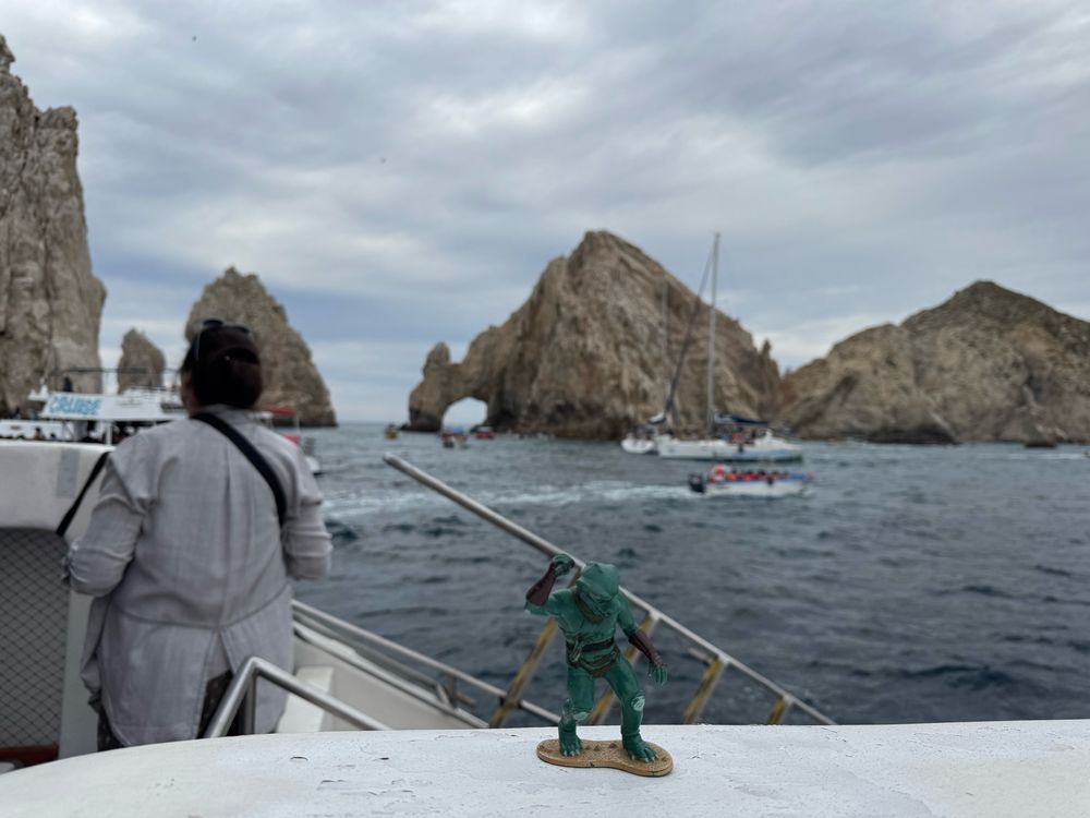 From the deck of a boat, the green lizard like Gorn action figure is posed in front of jagged rocks rising from the ocean. In the brown rocks is a single arch at the very tip of the Baja California peninsula. 