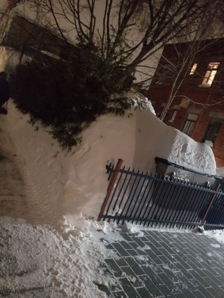 A photo of the corner of a fenced yard with snow piled up to twice the height of the gate. There is a mostly cleared sidewalk in the foreground.