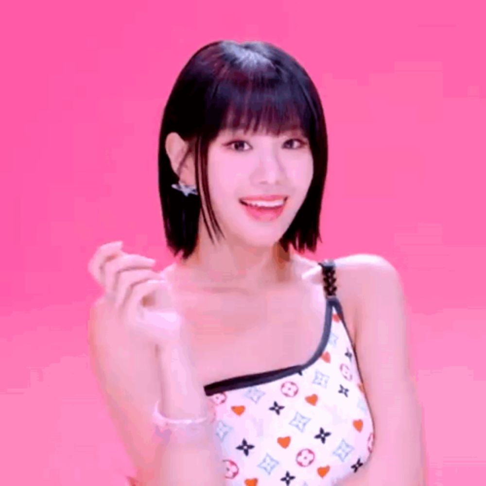 a woman in a one shoulder top is smiling and making a heart with her hand .