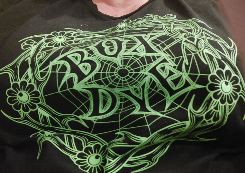 Black tshirt with intricate green design, a bit like a cobweb surrounded by a flower wreath. The band name 'box dye' is in the centre of the web. 
