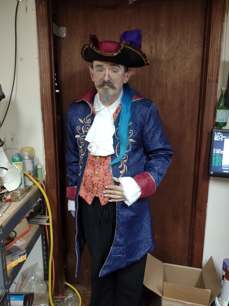A striking blue-haired goblin in full Regency regalia poses in a cluttered workshop. He wears a royal blue brocade coat with gold embroidery, a crimson brocade waistcoat, a white jabot, and a dramatic tricorn hat adorned with a purple plume. His hand rests on his hip with practiced elegance, radiating confidence, defiance, and impeccable style.
