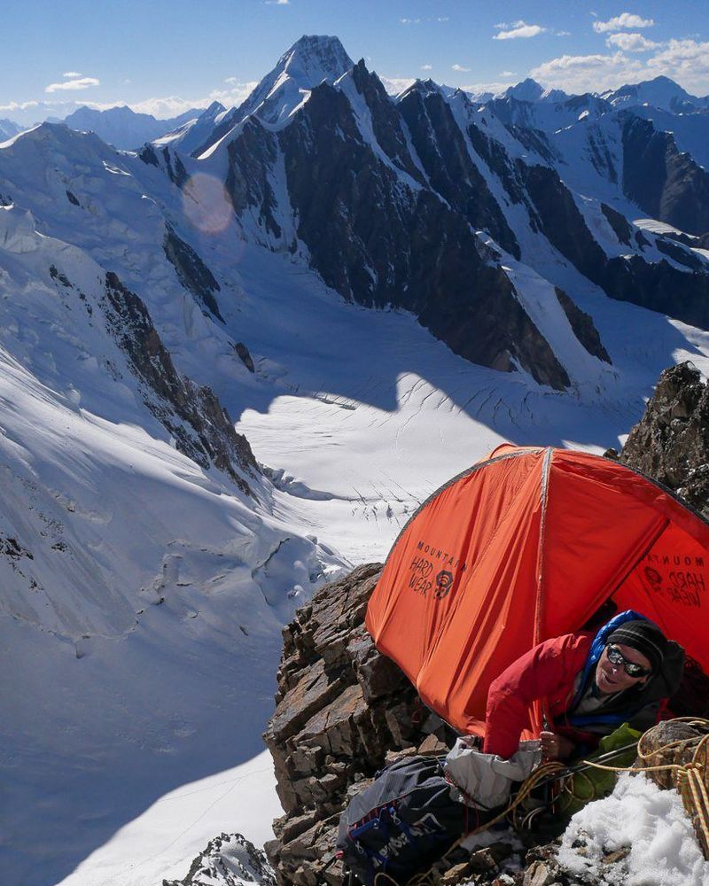 A climber pokes his head out of a small tent perched precariously on a rock promontary. Mountains and glaciers can be seen below him in the background.