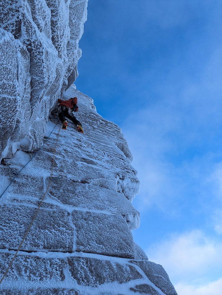 A climber pulling gear off their harness as they ascend a slabby hoar-frost-coated corner.