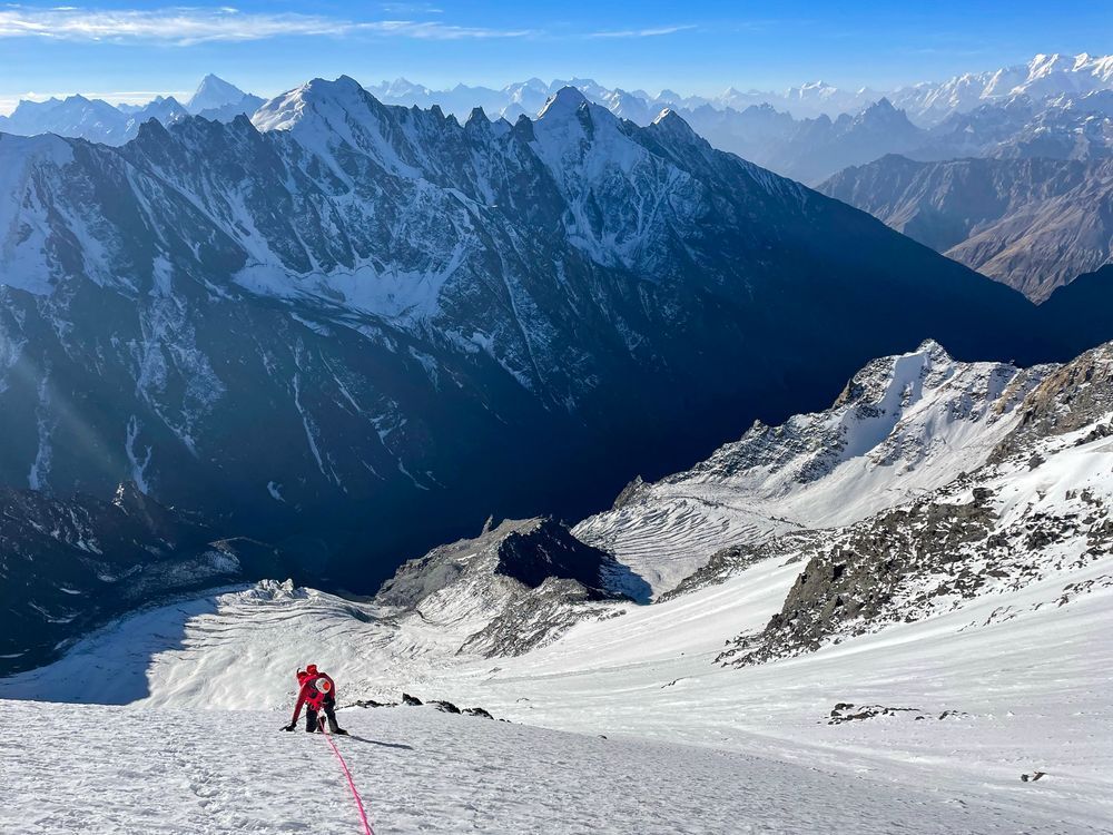 A climber in red ascends a snow slope, their pink rope snaking towards the camera. In the background a valley and series of rocky peaks are visible.