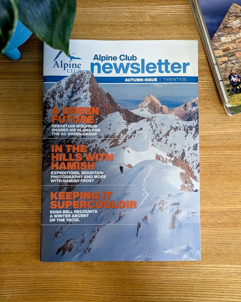 A newsletter (title: 'Alpine Club newsletter') lies flat ona  wooden table showing its front cover: a mountain ridge overwritten by section sub-headings. The newsletter is surrounded by a plant and an OS map to the Cairngorms.