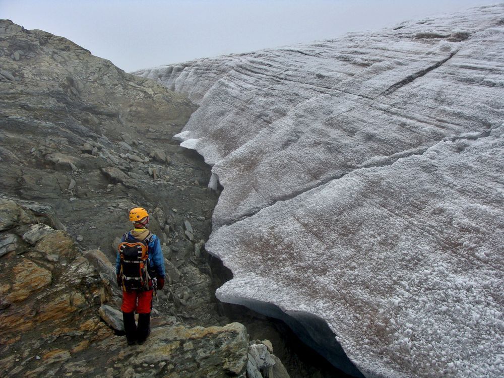 A figure in a backpack, harness and climbing helmet stands in front of the retreating edge of a glacier, inspecting the churned rock and earth left in its wake.