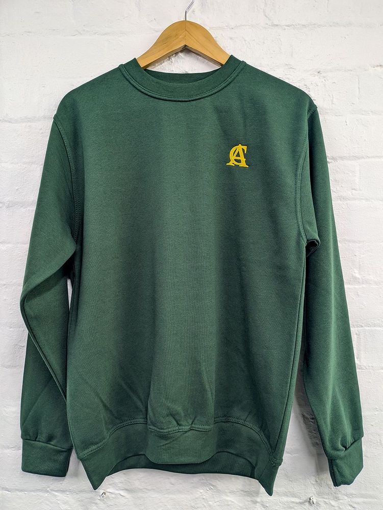 A green jumper with a gold 'AC' logo in front of a white brick wall.