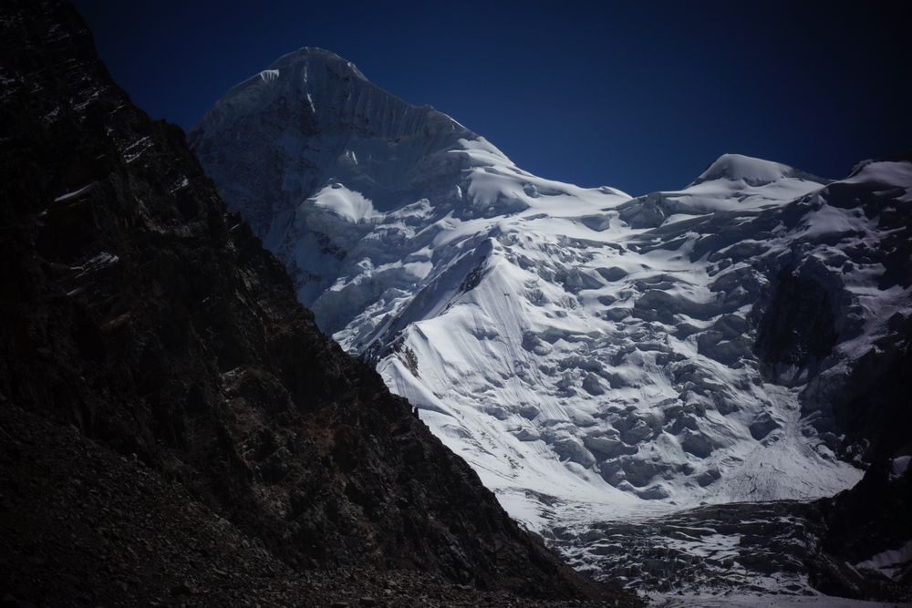 A mountain peak rises from a complex, broken glacier, a perfect snowy ridge rising to its peak in the left of the photograph.