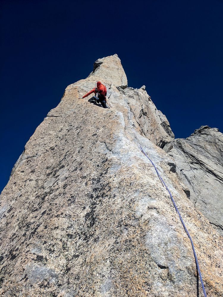 A climber ascending away from the camera up a pinnacle of rock framed against a dark blue sky.