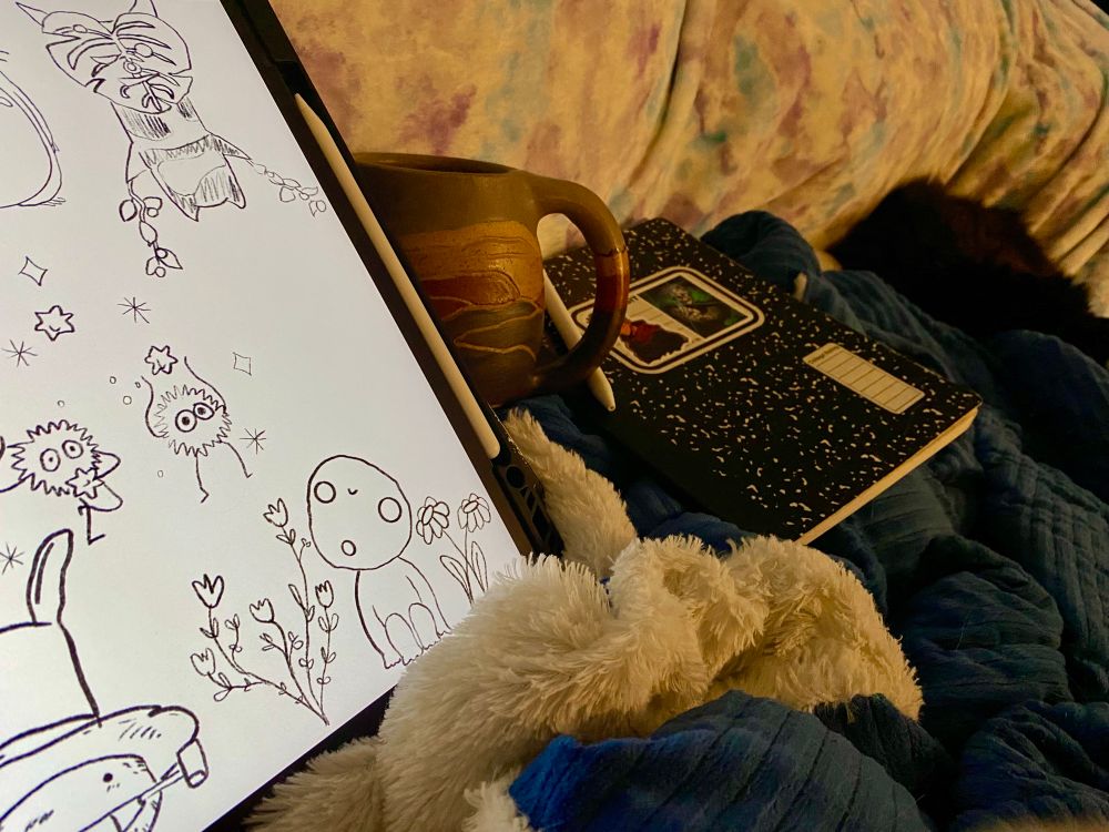 Photo of iPad with drawings, coffee mug, pen and notebook, and a cat on a blue textured blanket. Behind is a colorful throwblanket. 
