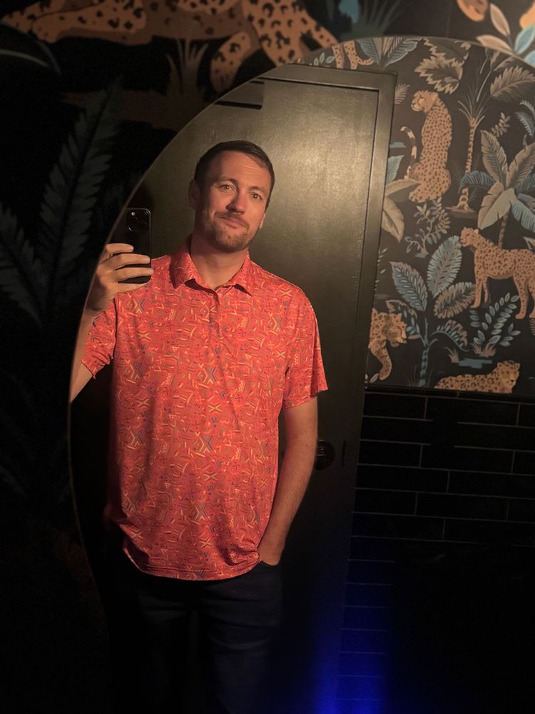 A tall, white guy with a beard and bearish smile takes a selfie in a bathroom mirror, wearing a pink patterned polo. The room is black trim and leopard and jungle themed wallpaper.  