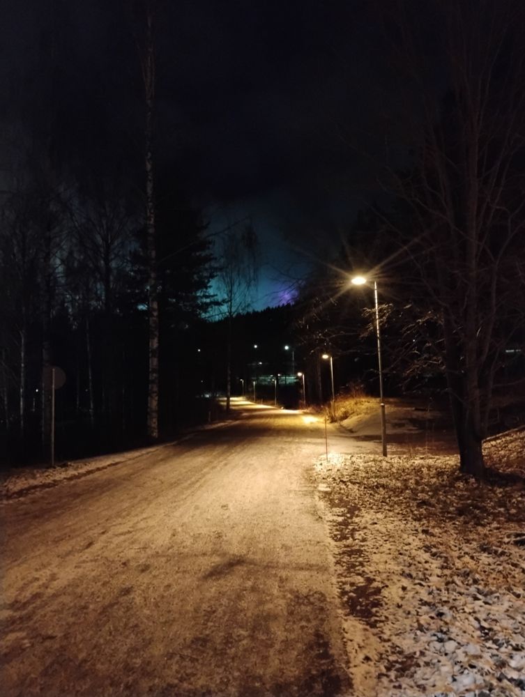 Ohut lumipeite pyörätiellä ja sen ulkopuolella. Puijontorni violettina taustalla pimeän metsän päällä.