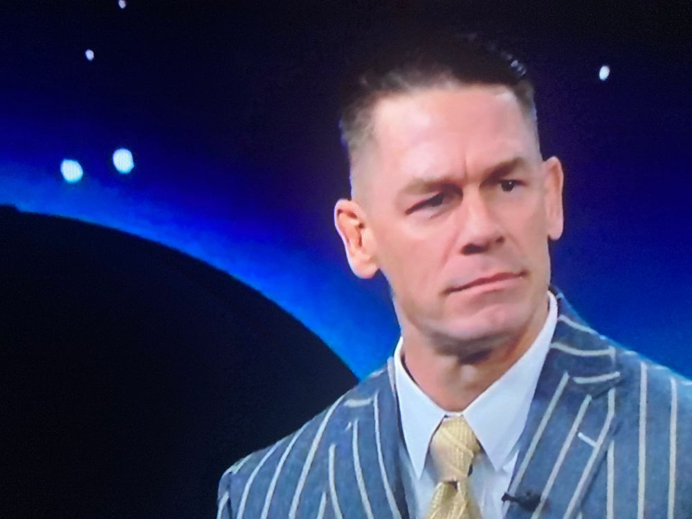 John Cena, cropped and coifed shiny brown hair, looking fuckin slick, swarthy, angular faced with his eyebrow raised like Spock from Star Trek as if to convey “This surprises me” in a bluish pinstripe suit, white collar shirt under and muted shiny gold tie.  Background is like black with a blue corona radiating from a broad black iris.